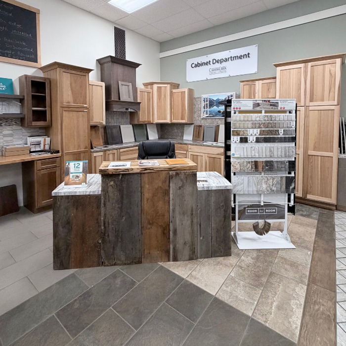 Interior of Carpet One flooring showroom in Eastern Oregon with multiple wood cabinet and panel displays, a central table with sample boards, and a wall sign reading “Carpet Department” above the displays.
